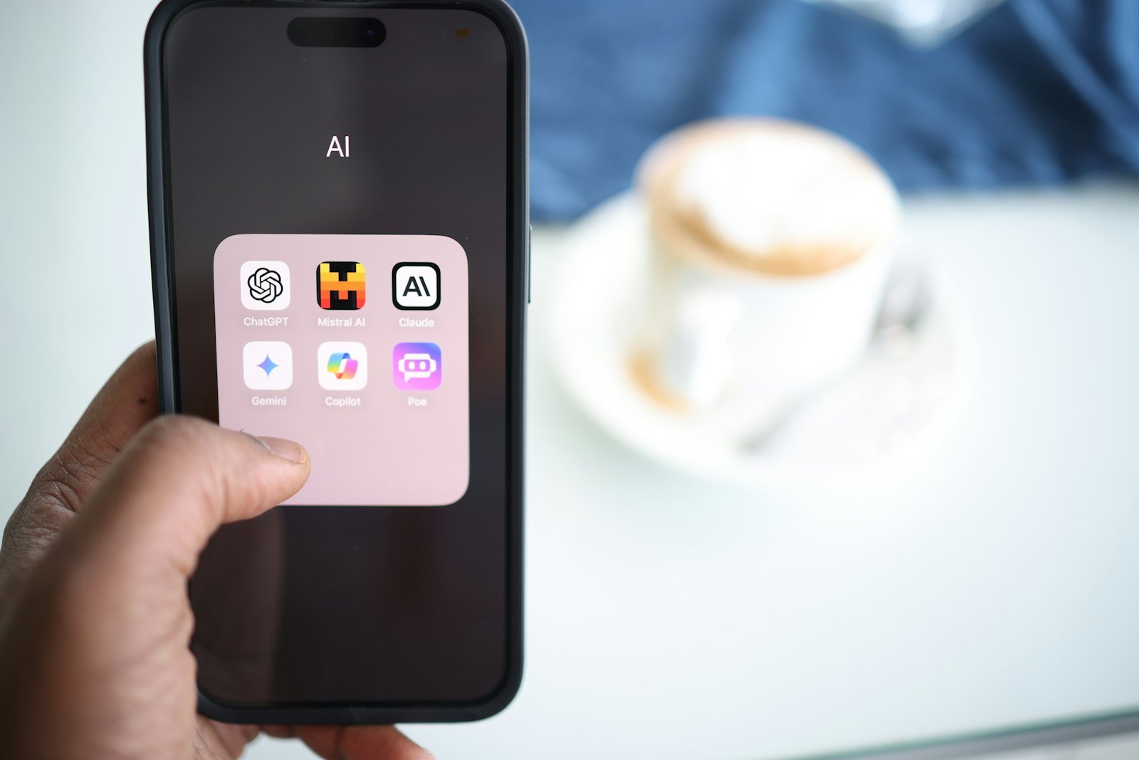 Hand holding a phone with a folder named AI containing assistant apps such as ChatGPT, Claude, Gemini and Copilot