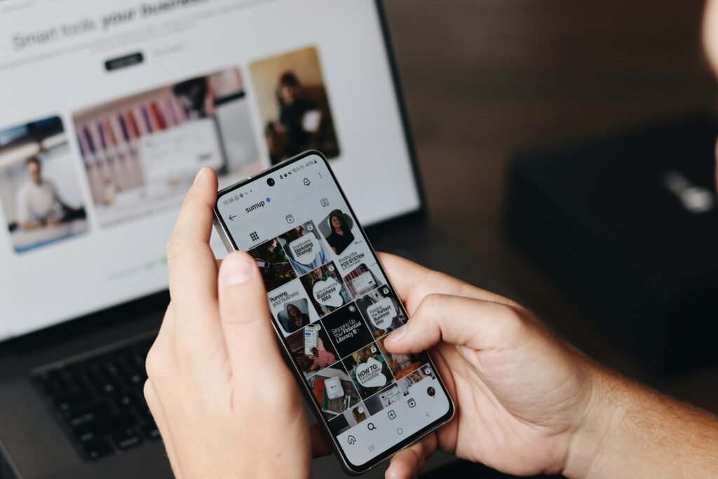 Hands holding a phone showing an Instagram feed in front of a laptop