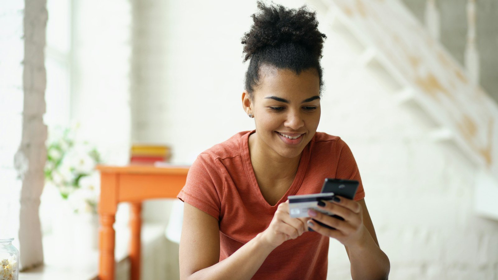 Smiling young woman holding a credit card and a phone while shopping online at home