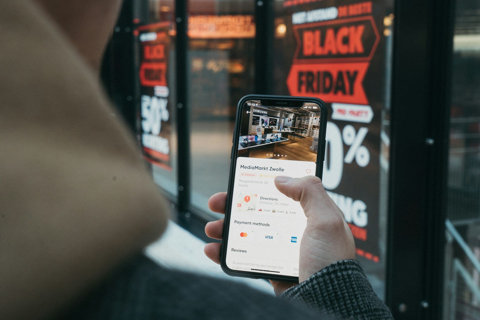 Shopper checking a phone in front of a store window covered in Black Friday sale signs