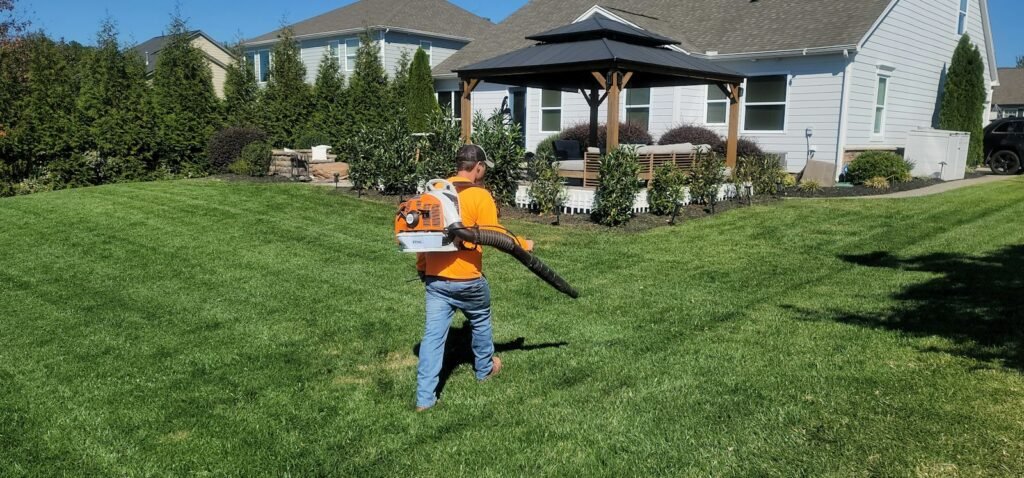 Yard service worker in an orange shirt clearing a lawn with a leaf blower in front of a house