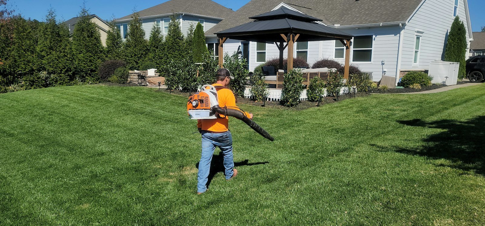 Yard service worker in an orange shirt clearing a lawn with a leaf blower in front of a house
